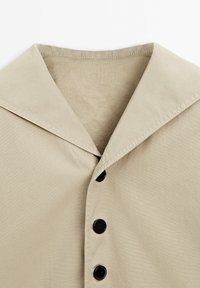 Chemise beige avec un large col plat et des boutons noirs sur le devant, fabriquée à partir d'un tissu lisse avec un léger éclat.