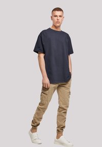 Jūrų laivyno medvilnės marškinėliai su trumpomis rankovėmis, derinami su smėlio spalvos kargo kelnėmis su šoniniais kišenėmis ir elastingais rankogaliais, užbaigti baltais sportiniais batais.