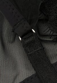 Svart nättyg med ett texturerat mönster, utrustat med justerbara remmar och en metallring. Sömnadsdetaljer är synliga på remmen.