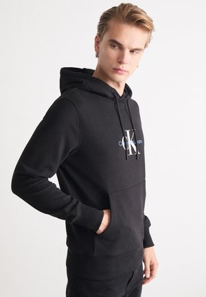 Sudadera negra con capucha, que presenta un bolsillo tipo canguro en la parte frontal, capucha ajustable con cordón y un logo en el pecho. Está fabricada con un tejido suave y cuenta con puños acanalados.