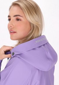 Violetinis nepralaidus vandeniui džemperis su dideliu gobtuvu, spaudžiamosiomis sagomis, pūkuotu pamušalu ir tekstiniu užrašu ant apykaklės. Lygus, blizgus paviršius.