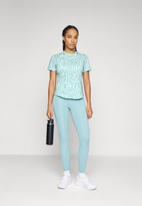 Tricou sportiv de culoare albastru deschis cu un model, asortat cu colanți. Persoana ține o sticlă de apă neagră și poartă adidași albi.