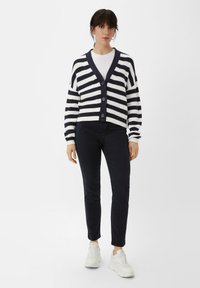Gestreifte Strickjacke in Marine und Weiß, mit V-Ausschnitt, Knopfverschluss und langen Ärmeln. Kombiniert mit dunklen Hosen und weißen Sneakers.
