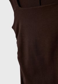 Gros plan sur un vêtement sans manches, brun foncé, avec un tissu lisse, légèrement ajusté et des coutures peu visibles sur un fond clair.