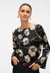 Mujer con el cabello recogido, lleva pendientes de aro dorados y una blusa negra con grandes flores beige, combinada con vaqueros oscuros y un cinturón.