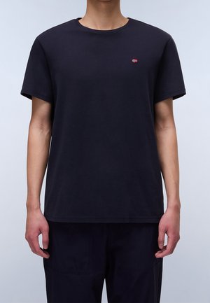 Person iført en ensfarvet sort kortærmet T-shirt med et lille norsk flaglogo på venstre bryst mod en lys baggrund.