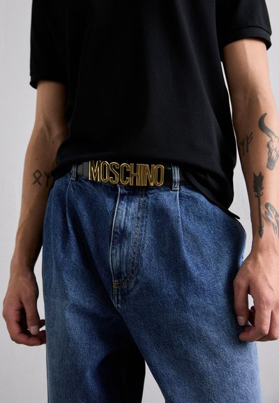 Čierne tričko skombinované s modrými džínsami. Zlato "MOSCHINO" opasok dopĺňa tento outfit. Viditeľné ruky, zobrazujúce tetovania.