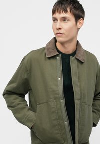 Maslinasto zelena jakna s rebrastim ovratnikom, prednjim kopčanjem na dugmad i dva velika zakrpa, nošena preko tamnozelene pletene vune.