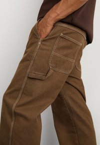 Pantalon cargo en coton marron avec poches latérales, détails de coutures blanches, et une coupe décontractée. Le tissu a une texture lisse et robuste.
