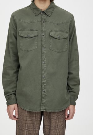 Mann trägt ein olivgrünes Hemd mit Knopfleiste und Taschen mit Klappen auf der Brust sowie braune karierte Hosen, steht vor einem schlichten Hintergrund.