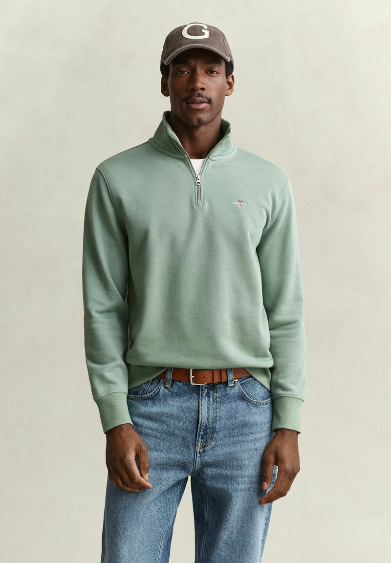 Man wearing light green quarter-zip sweatshirt, blue jeans with brown belt, and gray cap with white letter G on front.