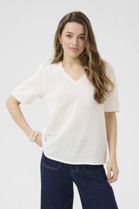 Blouse blanche texturée à manches courtes, décolleté en V et bordure en dentelle. Présente des points surélevés subtils sur l'ensemble. Portée avec un pantalon en denim bleu.
