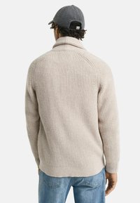 Man som bär en beige stickad tröja och blå jeans, vänd bort, med en grå keps, mot en vanlig vit bakgrund.