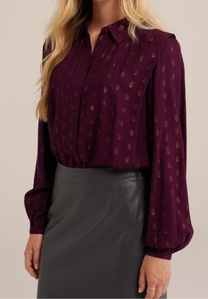 Woman wearing a long-sleeve purple blouse with metallic dots tucked into a high-waisted black leather skirt, standing against a neutral background.