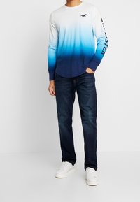 Långärmad tröja med gradient som övergår från vit till mörkblå, med en logotyp på bröstet och text på ärmen. Bärs med mörka jeans och vita skor.