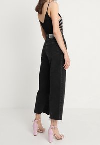 Jeans en denim noir taille haute avec des bords effilochés, associés à des sandales à talons hauts rose pâle. La taille est ornée d'une étiquette de marque.