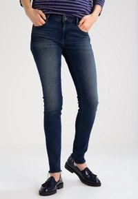 Jeans ajustados de color azul oscuro, hechos de denim con una textura suave, de cintura alta, y Diseño estándar de cinco bolsillos, combinados con zapatos negros de cordones.