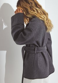 Grå uldblanding frakke med bælte i taljen, overdimensionerede ærmer og en buet kant. Har kontrastpipings for ekstra detalje.