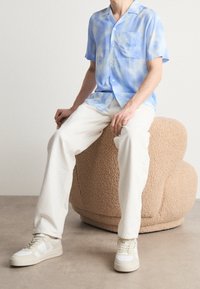 Ljusblå batikmönstrad kortärmad skjorta med knappar, krämfärgade löst sittande byxor och beige sneaktrar, sittande på en texturerad rund stol.