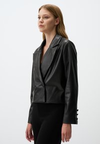 Zwarte leren cropped blazer met notch-revers. Beschikt over een dubbele knoopsluiting en drie knopen op elke manchette. Gladde textuur.