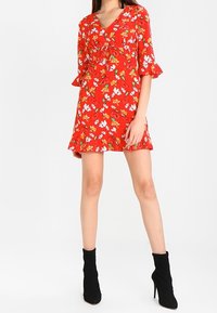 Rotes Blumenkleid mit kurzen, ausgestellt geschnittenen Ärmeln, Bindeband am Ausschnitt und Rüschensaum. Kombiniert mit schwarzen Stiefeletten.