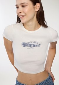 Biała krótka bluzka z krótkim rękawem typu crop top z niebieskim nadrukiem graficznym przedstawiającym samochód vintage oraz napis "SPEED QUEEN" poniżej; miękka tkanina o dopasowanym kroju.