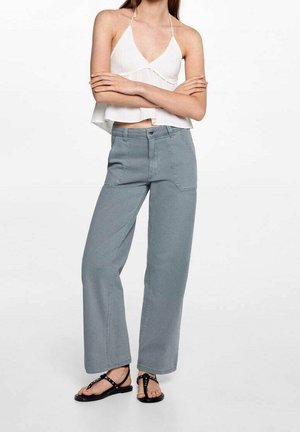 Nainen pukeutuneena valkoiseen halter-lyhytpaitaan, harmaisiin leveälahkeisiin housuihin, joissa on etutaskut, ja mustiin niitattuihin tasapohjaisiin sandaaleihin, seisomassa käsivarret ristissä.