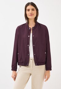 Vrouw in een paarse bomberjack over een witte top, met lichtgekleurde broeken, glimlachend tegen een witte achtergrond.