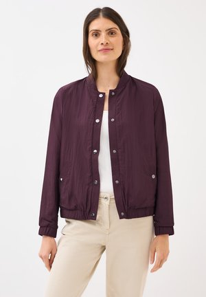 Woman wearing a purple bomber jacket over a white top, with light-colored pants, smiling against a white background.