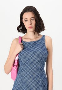 Denim obleka s modrim in belim kariranim vzorcem; enodelna zasnova z ramenom. Drži sijočo roza torbico z naramnico in kovinskimi detajli.
