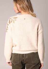 Crème-kleurige sweater met een losse pasvorm, ribbels aan de onderkant en een geborduurd bladerenmotief in goud op de linkerschouder. Zachte textuur.
