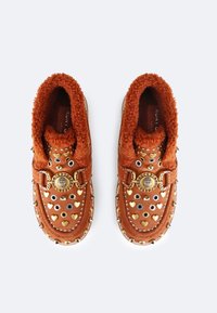 Brązowe zamszowe buty z puszystym pomarańczowym wykończeniem, ozdobami w kształcie serca i metalowymi ćwiekami, złotym wykończeniem oraz okrągłym emblematem z przodu.