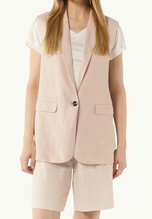 Femme portant un blazer sans manches rose pâle sur un t-shirt blanc, associé à un short assorti rose pâle arrivant aux genoux.