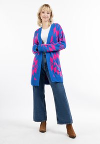 Modrý a ružový vzorovaný kardigán vyrobený z pleteného materiálu, s otvoreným predným dielom a dlhými rukávmi. V kombinácii s rozšírenými džínsami a hnedými členkovými čižmami.