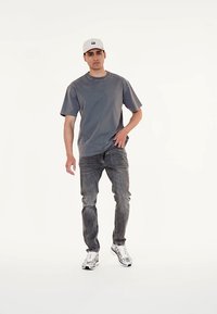 Camiseta de algodón gris de manga corta, combinada con jeans gris desgastados. El modelo lleva una gorra blanca y zapatillas plateadas. Diseño sencillo, sin patrones.