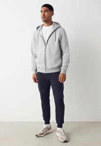 Sudadera gris con cremallera y capucha, combinada con pantalones jogger azul marino. Zapatillas beige y blancas. El tejido parece suave y texturizado.