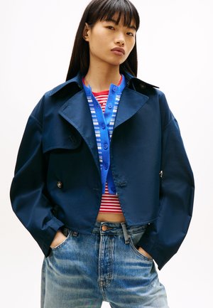 Młoda kobieta o długich ciemnych włosach nosi granatową krótka kurtkę na niebieskim kardiganie w paski oraz czerwonym topie w paski, w połączeniu z jasnoniebieskimi dżinsami.