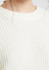 Maglione bianco lavorato a maglia con una texture a coste e un collo rotondo. Il tessuto appare morbido e accogliente con una leggera lucentezza.