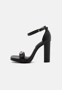 Svart høyhælte sandaler med tykk blokkhelt, ankelrem og en frontrem med metallbeslag. Glatt imitert skinntekstur.