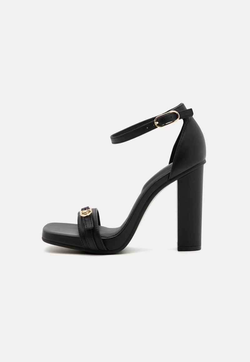 Svart høyhælte sandaler med tykk blokkhelt, ankelrem og en frontrem med metallbeslag. Glatt imitert skinntekstur.