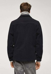 Chaqueta azul marino de mezcla de lana con cuello estructurado, ribete de punto texturizado y detalles de botones en las mangas. Combinada con vaqueros grises visibles en la cintura.