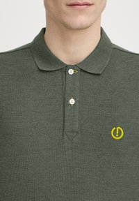 Mørkegrøn polo skjorte lavet af tekstureret stof, med krave, tre-knappet lukning og en gul logoaccent på venstre side.