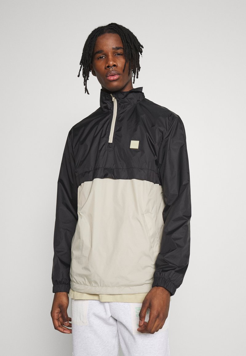 Giacca a vento nera e beige con colletto alto, mezza zip, polsini elasticizzati e una piccola patch con logo sul petto. Texture liscia.