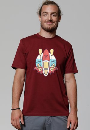 Man met baard en knot, gekleed in een bordeauxrode t-shirt met een kleurrijke afbeelding van een kajak en peddels op de voorkant.