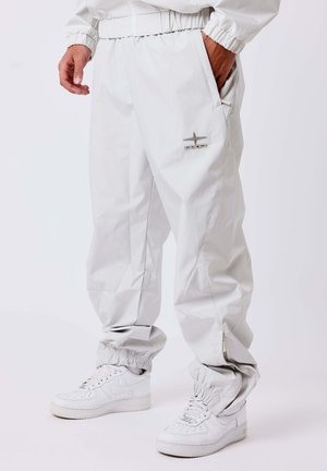 Parte inferior del cuerpo con pantalones deportivos blancos y holgados con puños elásticos y zapatillas blancas, una mano en el bolsillo del pantalón.