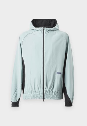 Gaiši zila vēja jakas ar melniem akcentiem. Iespējas iekļauj kapuci, rāvējslēdzēja priekšpusi, elastīgas aproces un savilktu apakšmalu. Materiāls izskatās gluds.