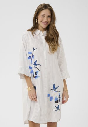 Žena oblečená v bielych šatách na zapínanie s jemnými pruhmi, zdobených modrým vtáčím a listovým vyšívaním, s pozlátenými kruhovými náušnicami a náramkami.