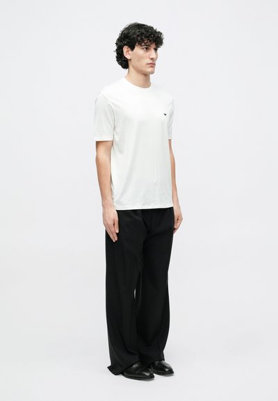 Fehér, rövid ujjú, kerek nyakú póló, bal mellrészen kis fekete logóval, fekete, bőszárú nadrággal és fekete cipővel párosítva.