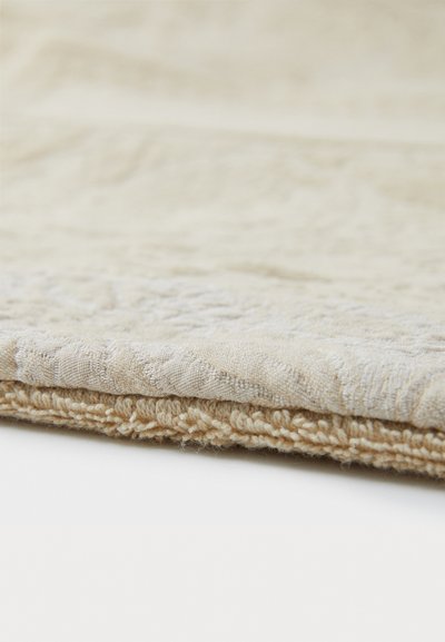 Beige textile with a textured surface and woven pattern along the edge. The fabric appears soft with a slight sheen.