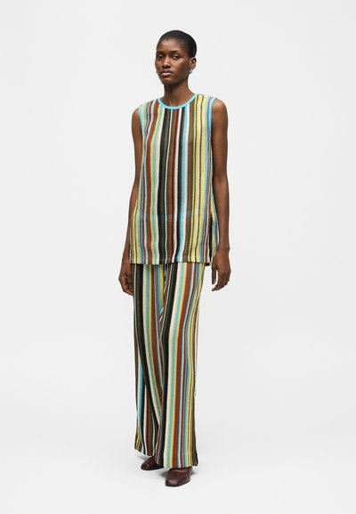Γυναίκα που φοράει αμάνικη μπλούζα με πολύχρωμες κάθετες ρίγες και ταιριαστό παντελόνι φαρδιάς γραμμής, στέκεται με τα χέρια χαλαρά στο πλάι.